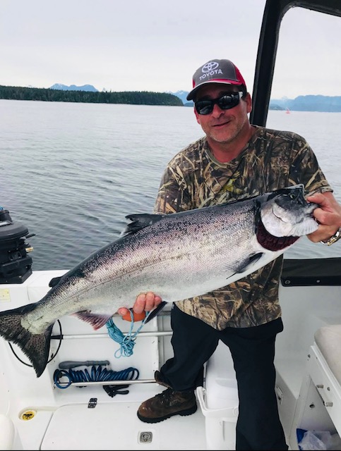 Douglas Smith, captain and guide of Kwigwis Adventures, smiles while holding a large silver salmon on his fishing boat. The boat deck is visible with fishing equipment nearby.